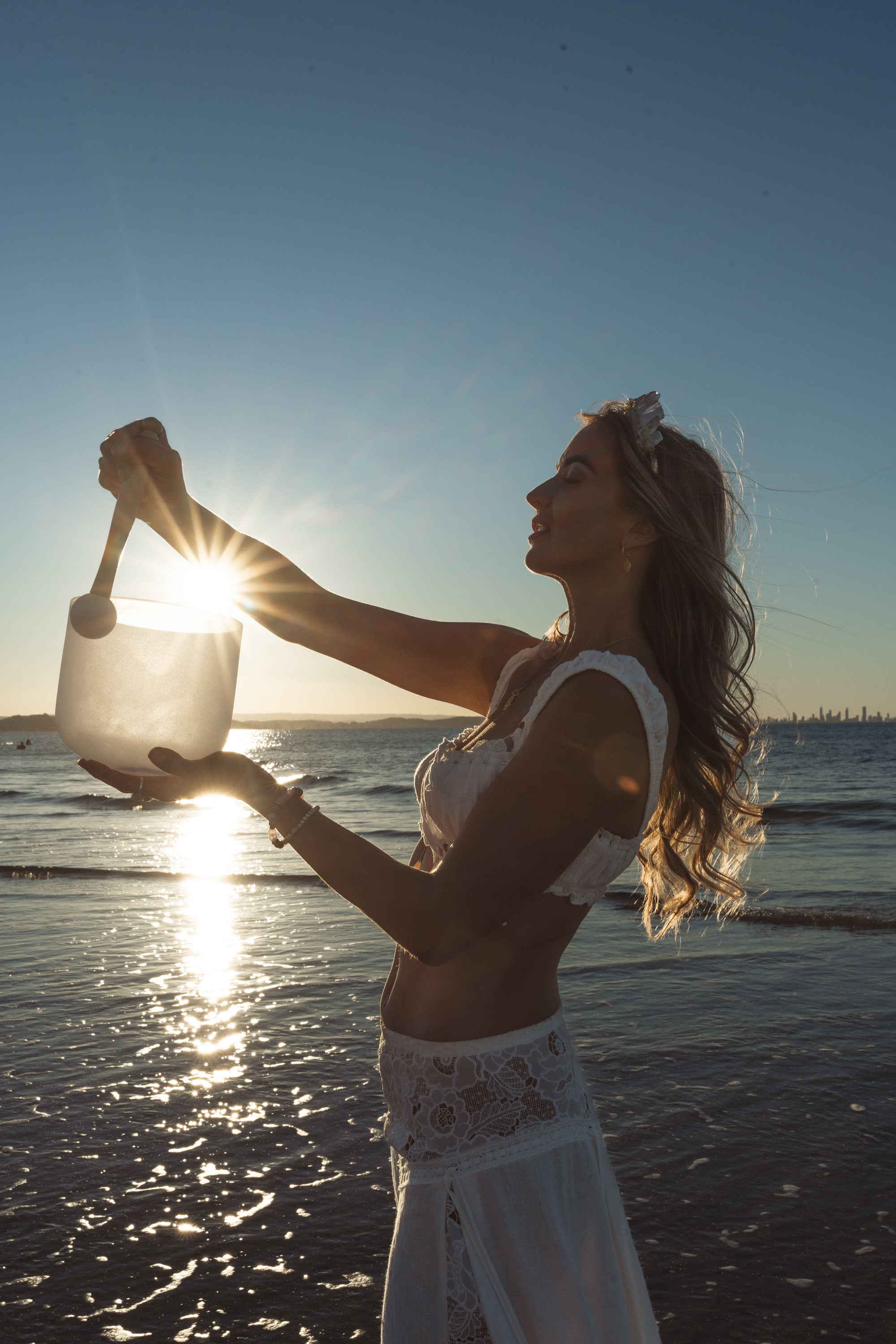 Bryony lifting a crystal singing bowl into the setting sun at the water's edge