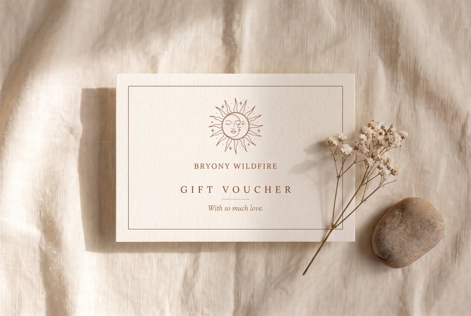 Bryony Wildfire gift voucher — a cream card with the sun and moon mark and the words 'With so much love', resting on ivory linen beside dried flowers and a river stone